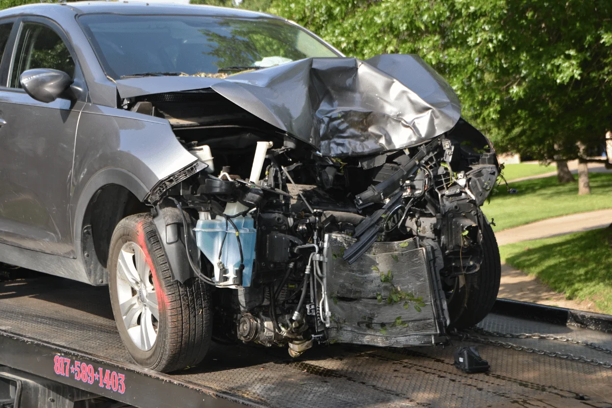 A silver car with severe front-end damage is on a tow truck, with its hood crumpled and parts exposed.