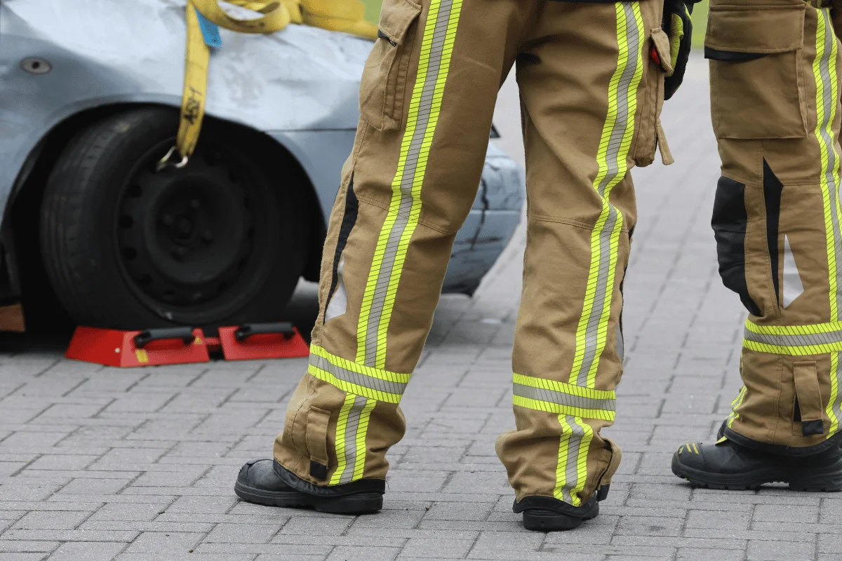 Firefighters in brown uniforms with reflective stripes stand on a paved road, near a damaged car wheel.