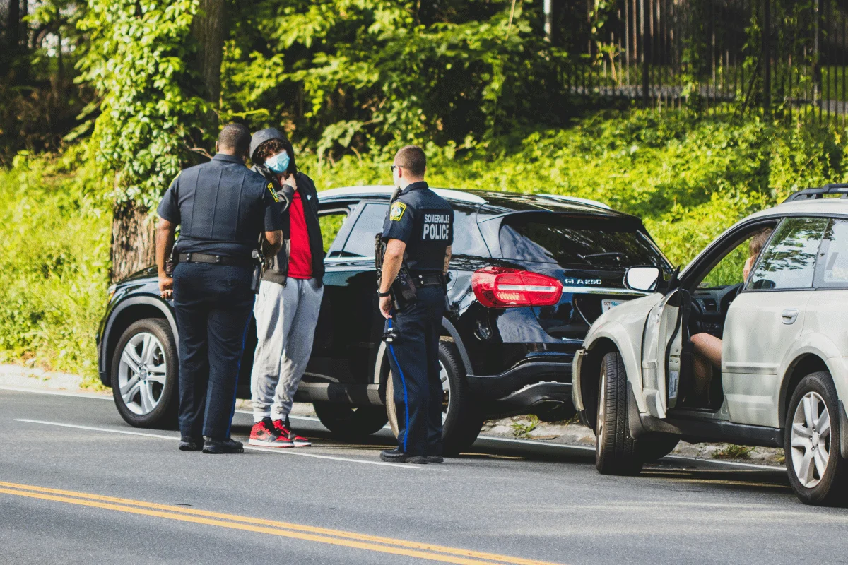 Two police officers stand beside a parked black car on a sunny road, engaging with a person wearing a mask and red jacket.