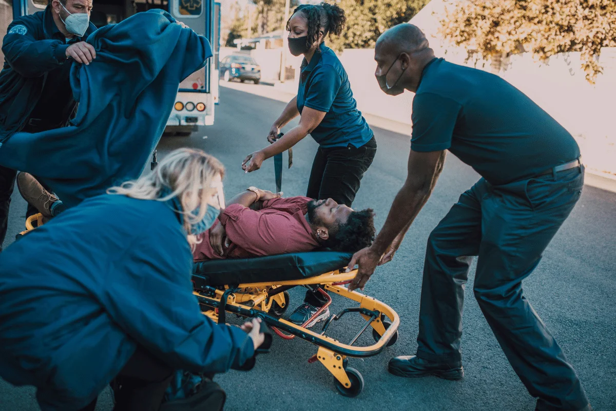 Emergency responders assist a person on a stretcher, wearing masks, on a sunny street.